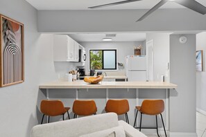 Eine moderne Küche mit weißen Schränken, einer zentralen Kochinsel mit Stühlen, einem Deckenventilator und Blick ins Freie.