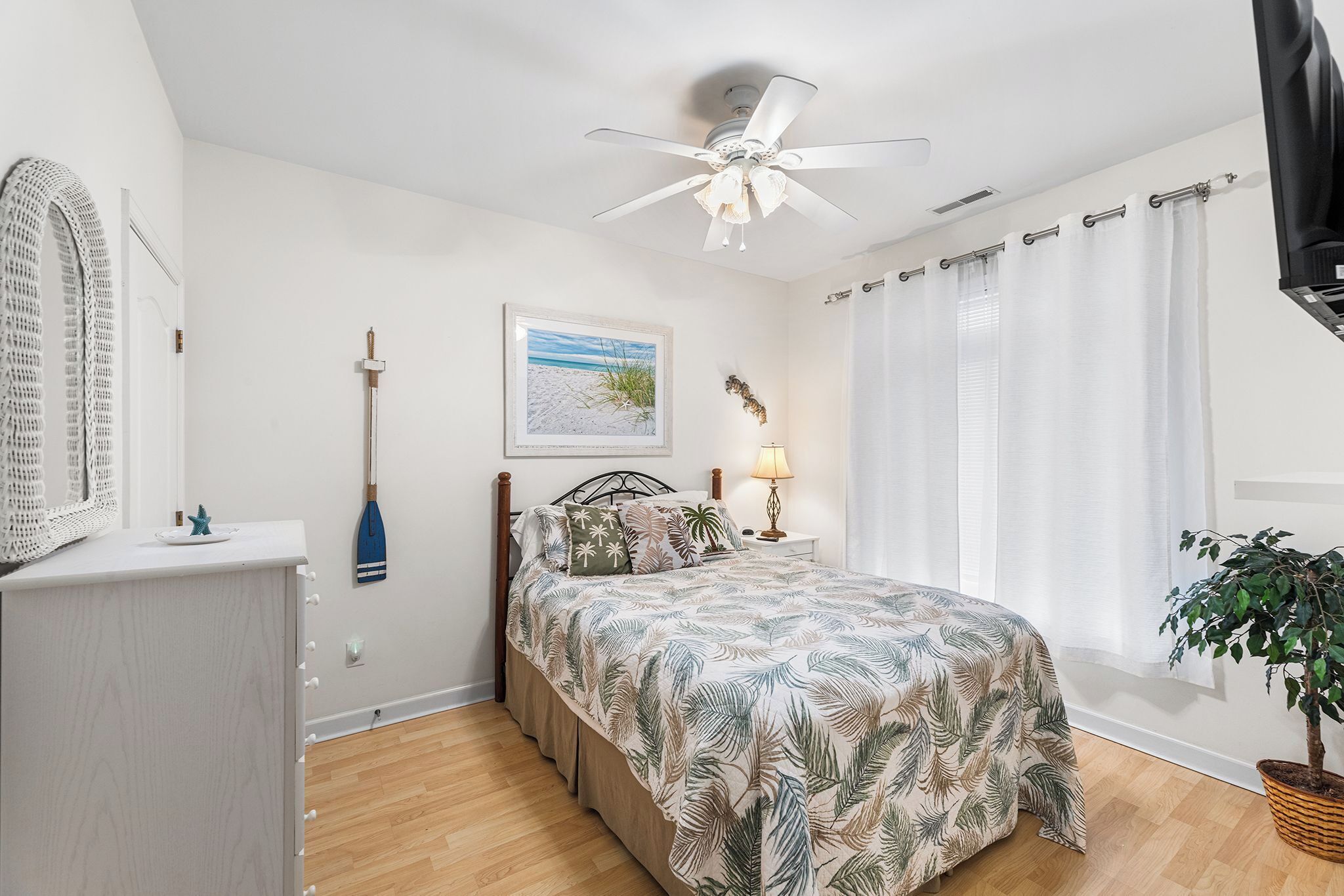 Une chambre à coucher avec un lit, un ventilateur de plafond, une fenêtre avec des rideaux, une lampe de chevet, une plante fixée au mur et un tableau encadré.