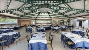 An indoor dining area with tables set for multiple people, a high ceiling with green structural beams, and a view of the outdoors. - Marina Living Rooms – Economy Double Room with Shared Pool, Wi-Fi & Air Conditioning (Laura)