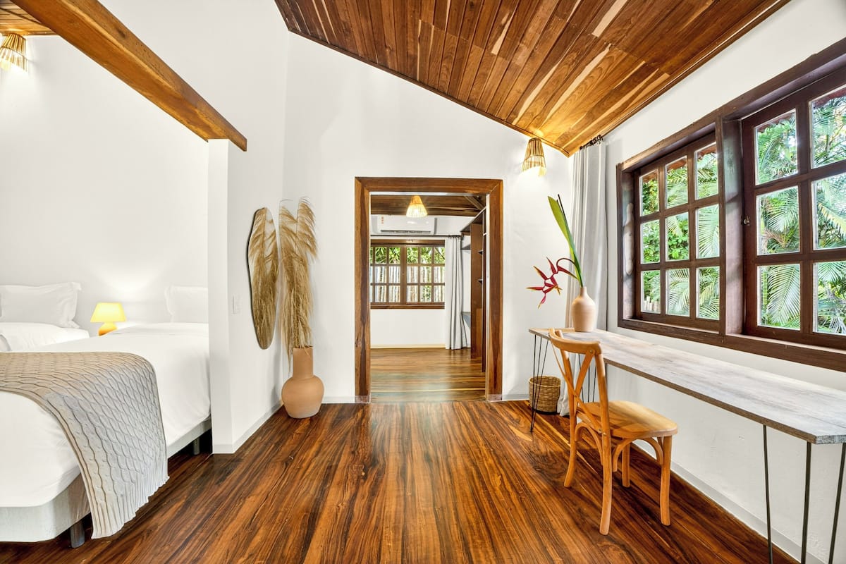 A room with a bed, a wooden ceiling, a window with a view, and a desk with a chair.