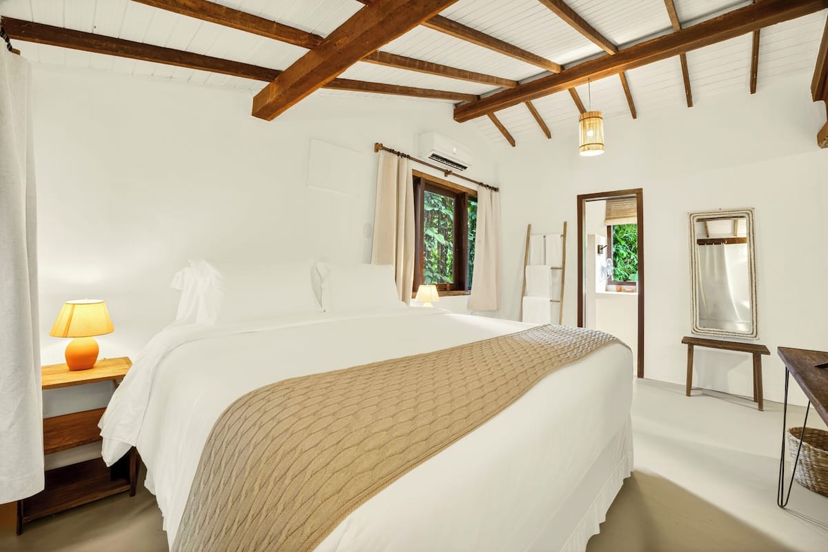 A bedroom with a large bed, a bedside table with a lamp, a window with curtains, a mirror, and a wooden ceiling.