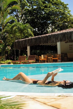 A pool area with lounge chairs, a thatched-roof structure, and lush greenery. - Vila Luna (Porto Seguro)