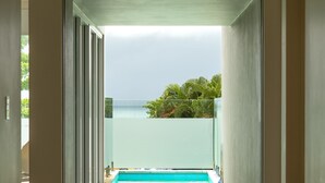 On aperçoit, à travers une porte vitrée, une piscine, avec une vue dégagée sur l’océan et les palmiers.