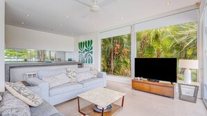 Un salon moderne avec un canapé gris, une table basse, un téléviseur et une grande fenêtre donnant sur une végétation tropicale.