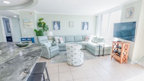 A modern living room with a curved marble countertop, a sectional sofa, a round ottoman, and a television on a wooden stand. - 1815 North Hampton, Ocean View Condo With Full Access!! 3 Bedroom Condo (Myrtle Beach)