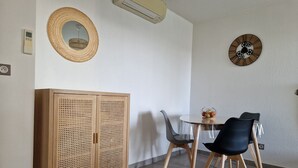 Zona pranzo con tavolo rotondo, due sedie, un mobile con ante forate, condizionatore d'aria a muro, orologio a parete e presa elettrica.