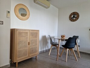 Un coin repas comprenant une table ronde, deux chaises, un meuble avec des portes perforées, un climatiseur fixé au mur, une horloge murale et une prise de courant.