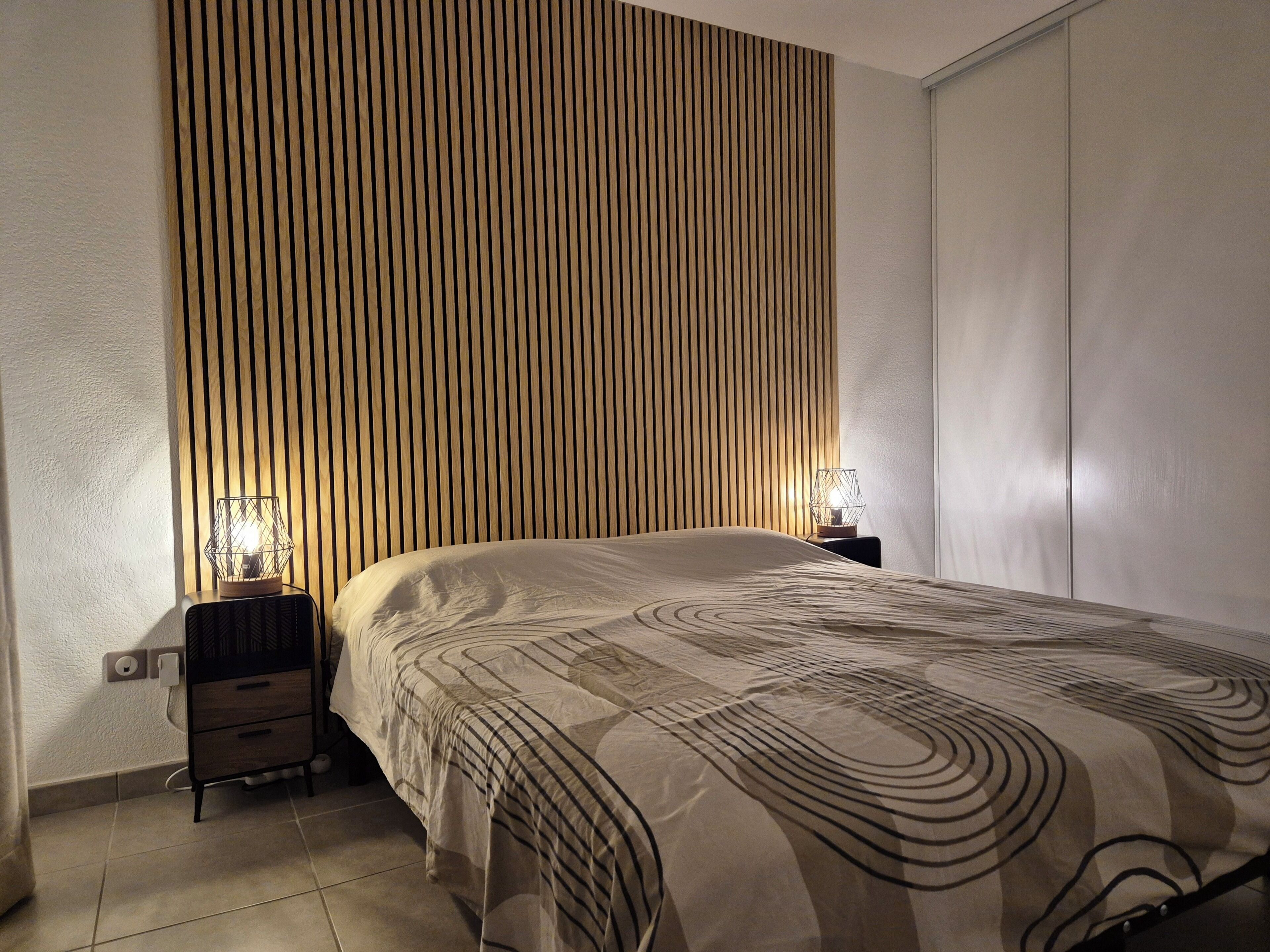 A bedroom with a bed, two bedside tables, a wall with vertical wooden slats, and a nightstand with a lamp.