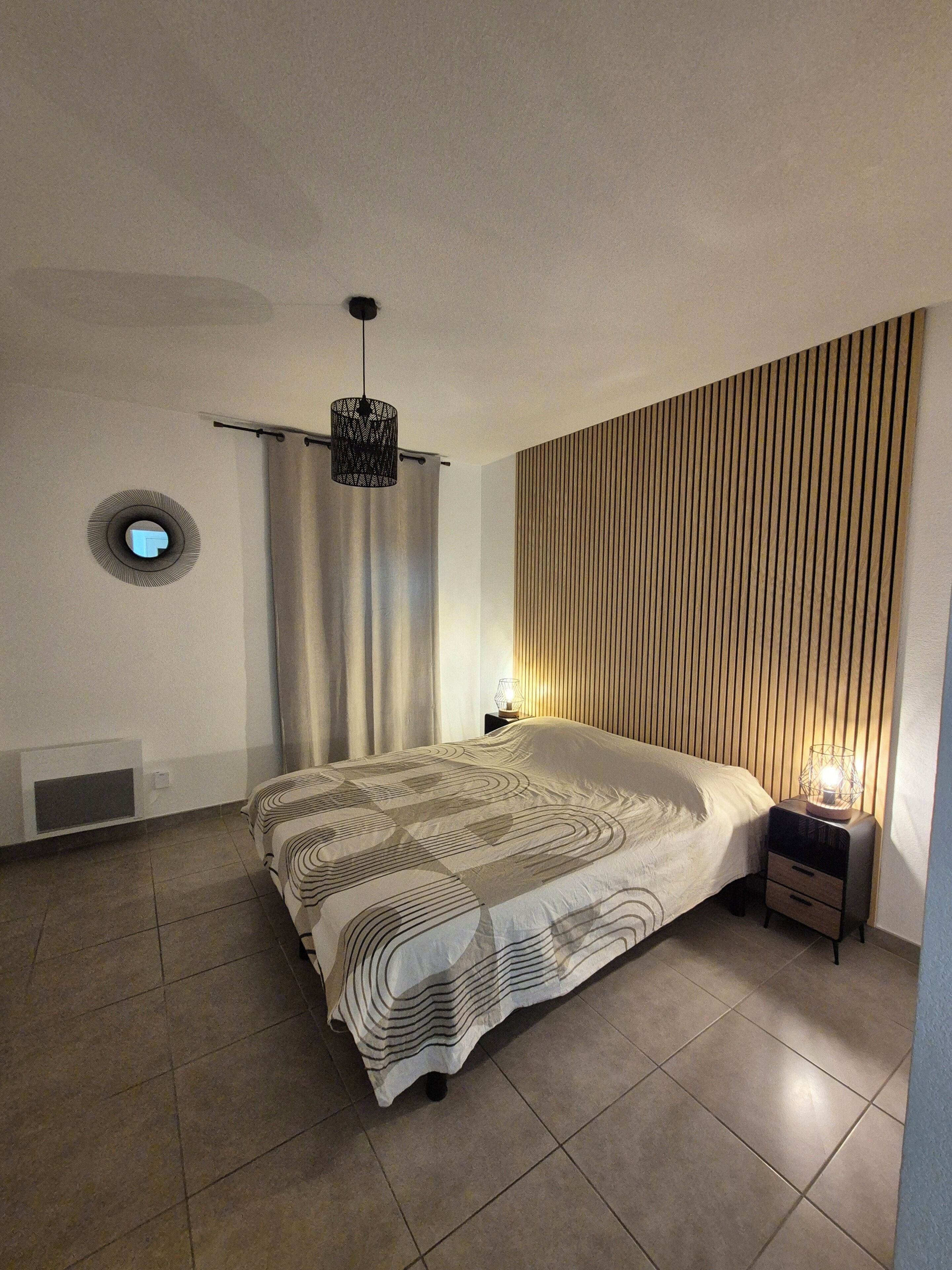 A bedroom with a bed, a nightstand, a wall-mounted light, a ceiling vent, and a curtain.