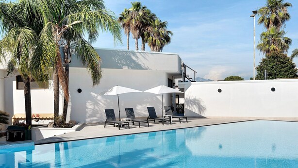 A swimming pool with lounge chairs and umbrellas, surrounded by palm trees and a white building. - Marina Living Rooms – Economy Double Room with Shared Pool, Wi-Fi & Air Conditioning (Laura)