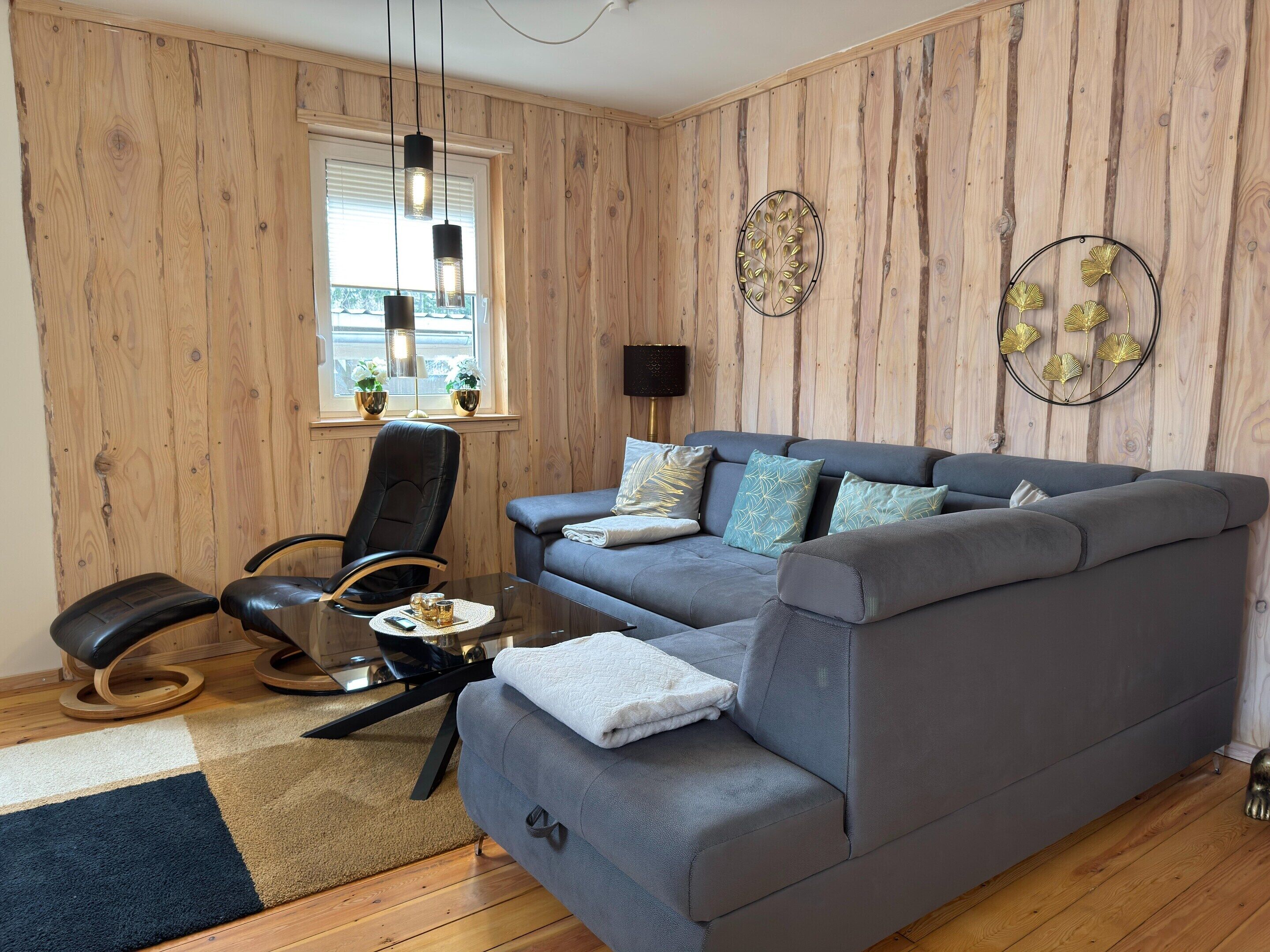 Una sala de estar moderna con un sofá seccional gris, un sillón de cuero negro, una mesa de centro de vidrio y paneles de madera.