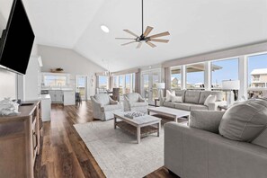 A spacious living area with a large window, a ceiling fan, and a kitchen in the background. - Pier Pressure - 301 Port Royal St 3 Bedroom Home (Dauphin Island)