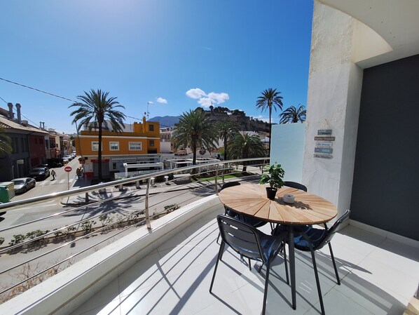 Ein Balkon mit rundem Tisch und Stühlen, Blick auf eine Straße mit Autos und Palmen, im Hintergrund Gebäude.