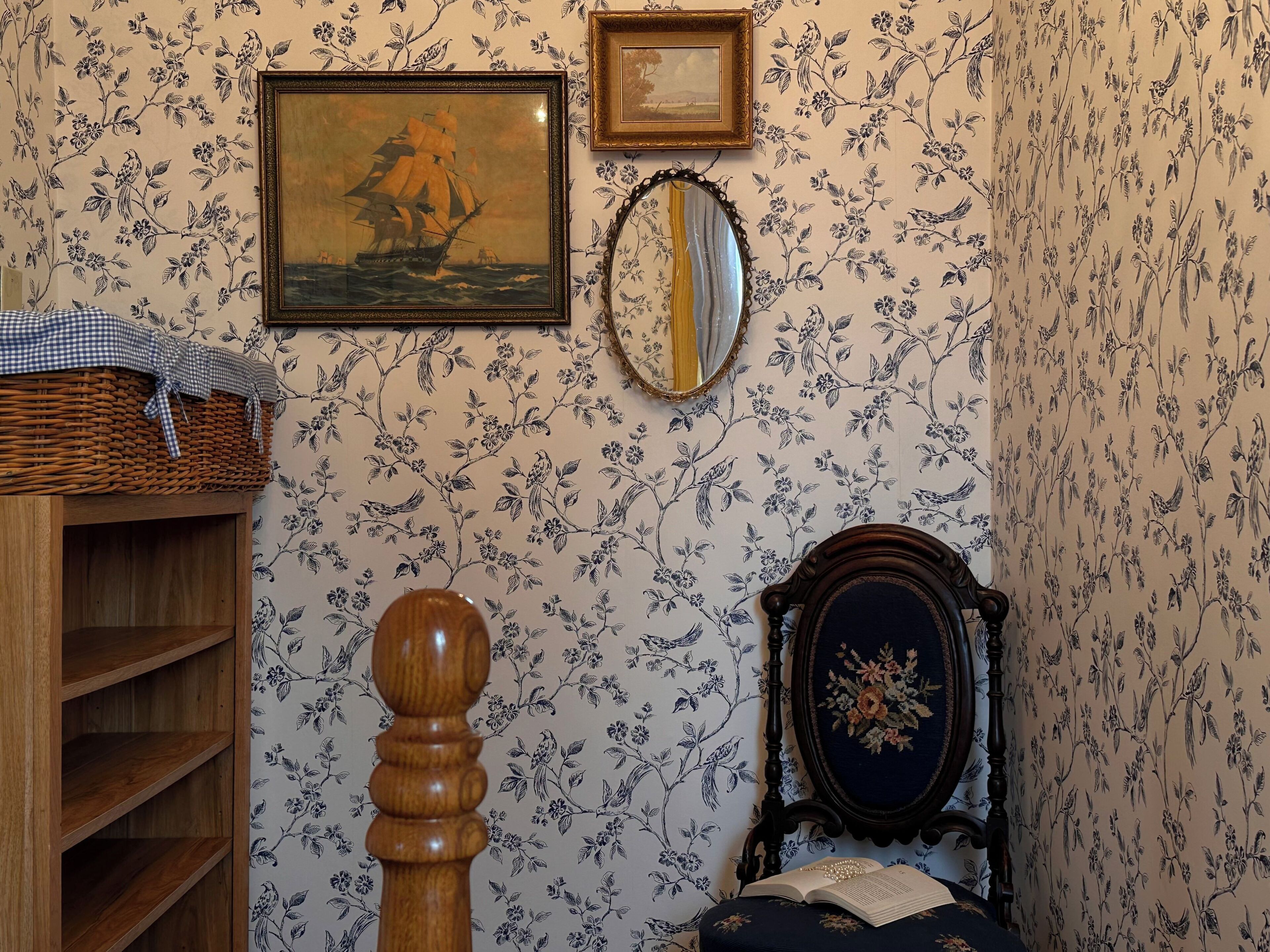 Habitació amb una prestatgeria de fusta, un quadre amb un vaixell, un mirall i un llit amb un estovat floral.