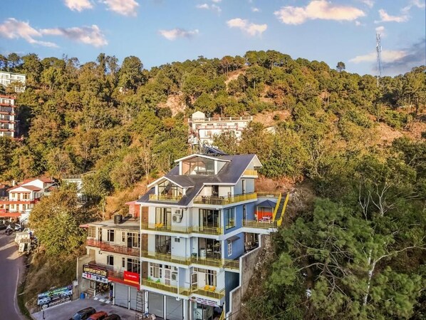 A multi-story hotel with balconies, a parking area, and a view of a hillside. - Hillshades Retreat by Alaya Stays 6BHK (Solan)