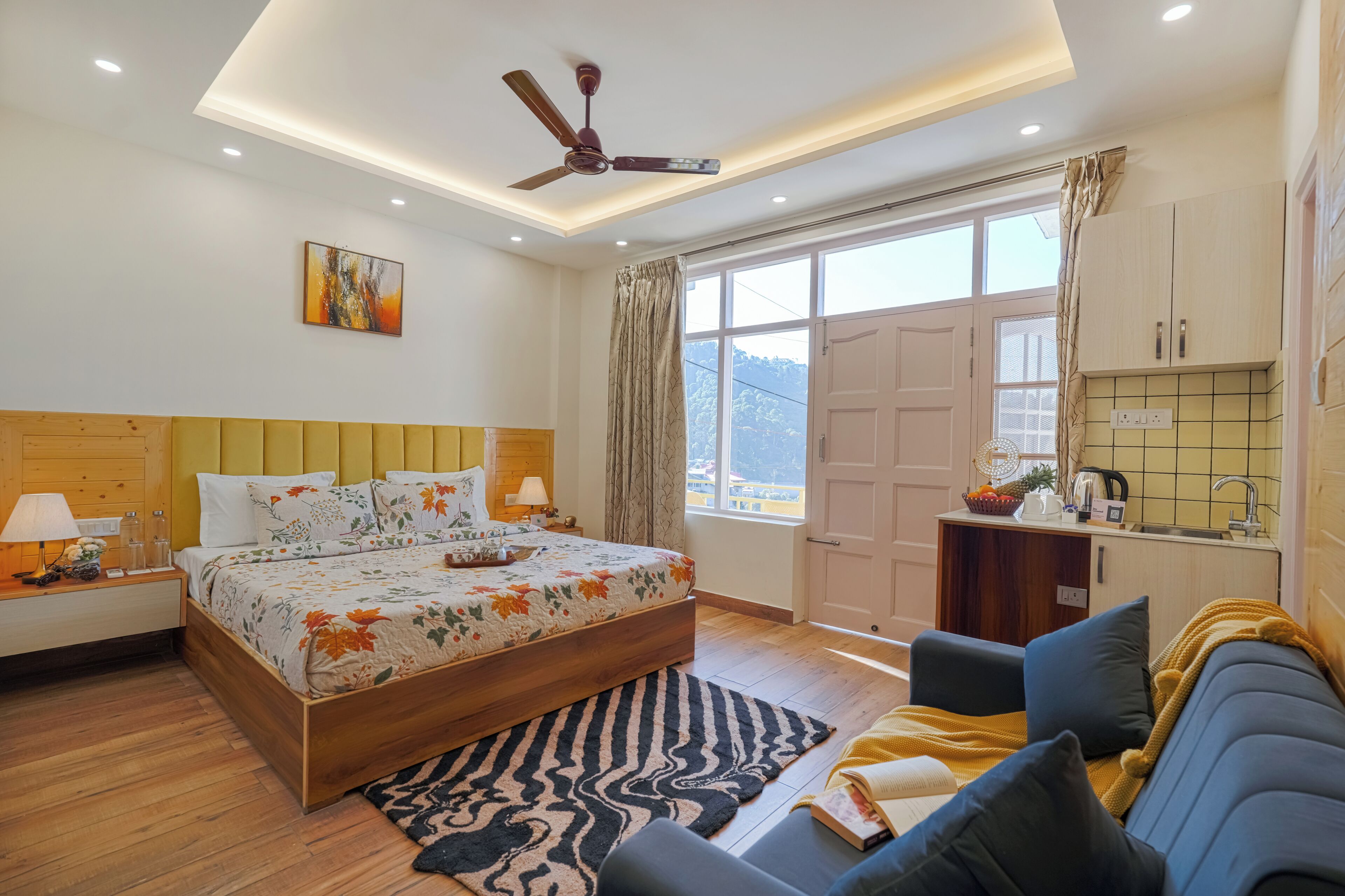 A bedroom with a wooden bed, a ceiling fan, a nightstand, a bedside table, a sofa, a window with curtains, and a view of the outdoors.