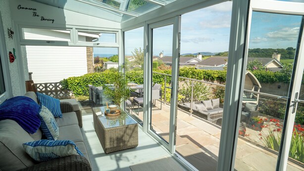 Ein sonnendurchflutetes Zimmer mit einer Couch, einem Korbtisch und einer Glastür zu einer Terrasse mit Gartenmöbeln und Blick auf die Nachbarschaft.