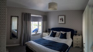 Ein Schlafzimmer mit Bett, einem Fenster mit Vorhängen, einem Nachttisch, einer Lampe und einem gerahmten Bild an der Wand.