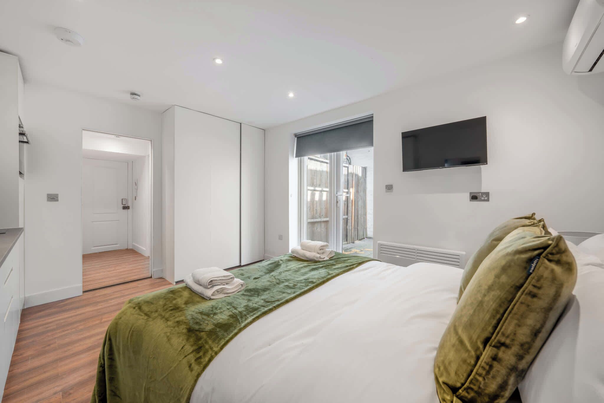 A modern hotel room with a bed, a green throw, a flat-screen TV, and a sliding door leading to a balcony.
