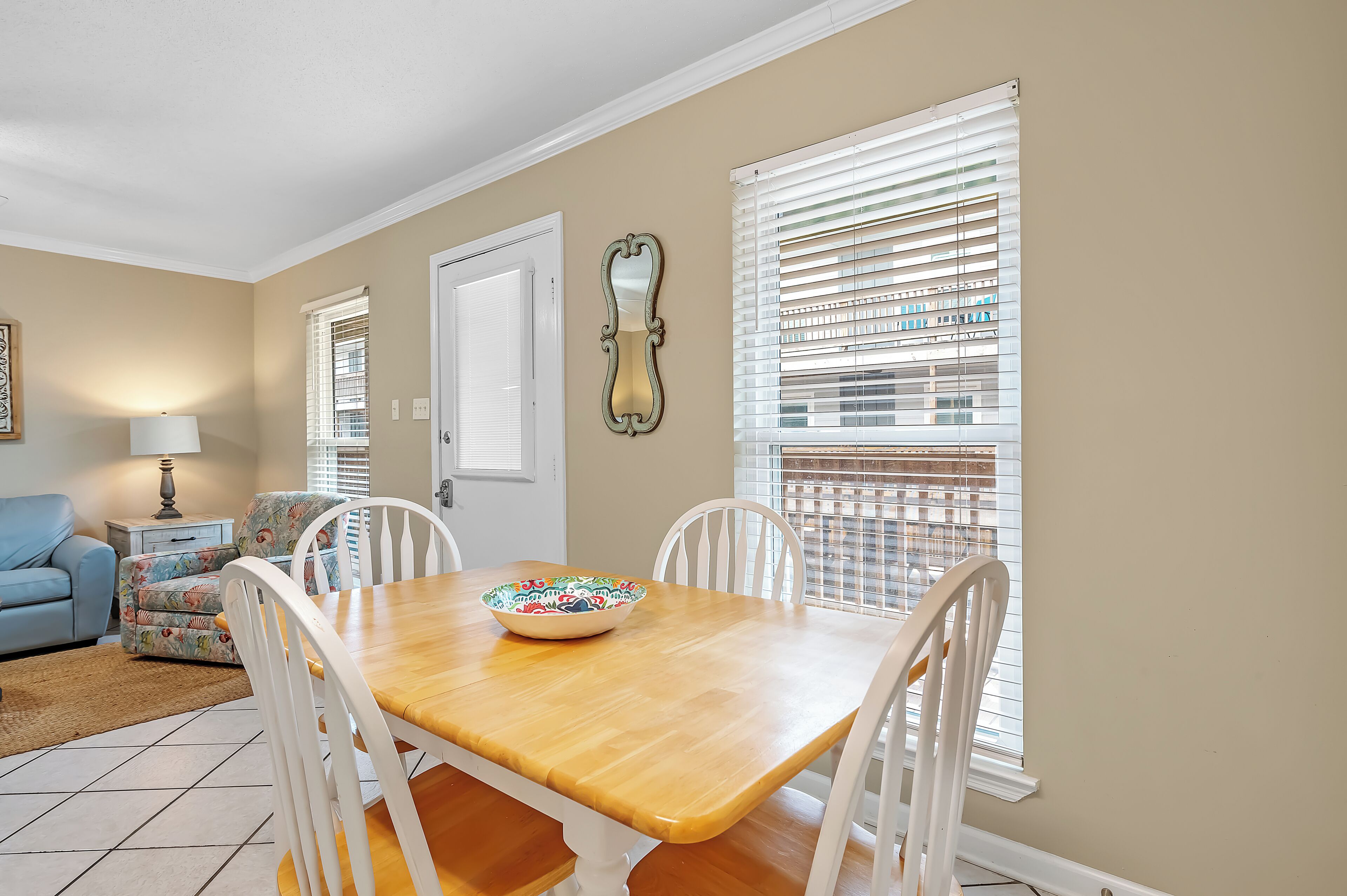 A dining area with a wooden table, white chairs, a colorful bowl, a lamp, and a window with blinds.