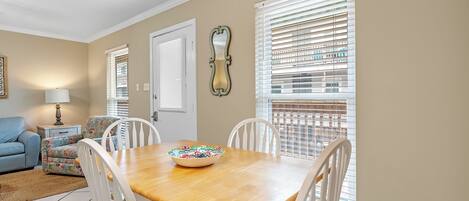 A dining area with a wooden table, white chairs, a colorful bowl, a lamp, and a window with blinds.