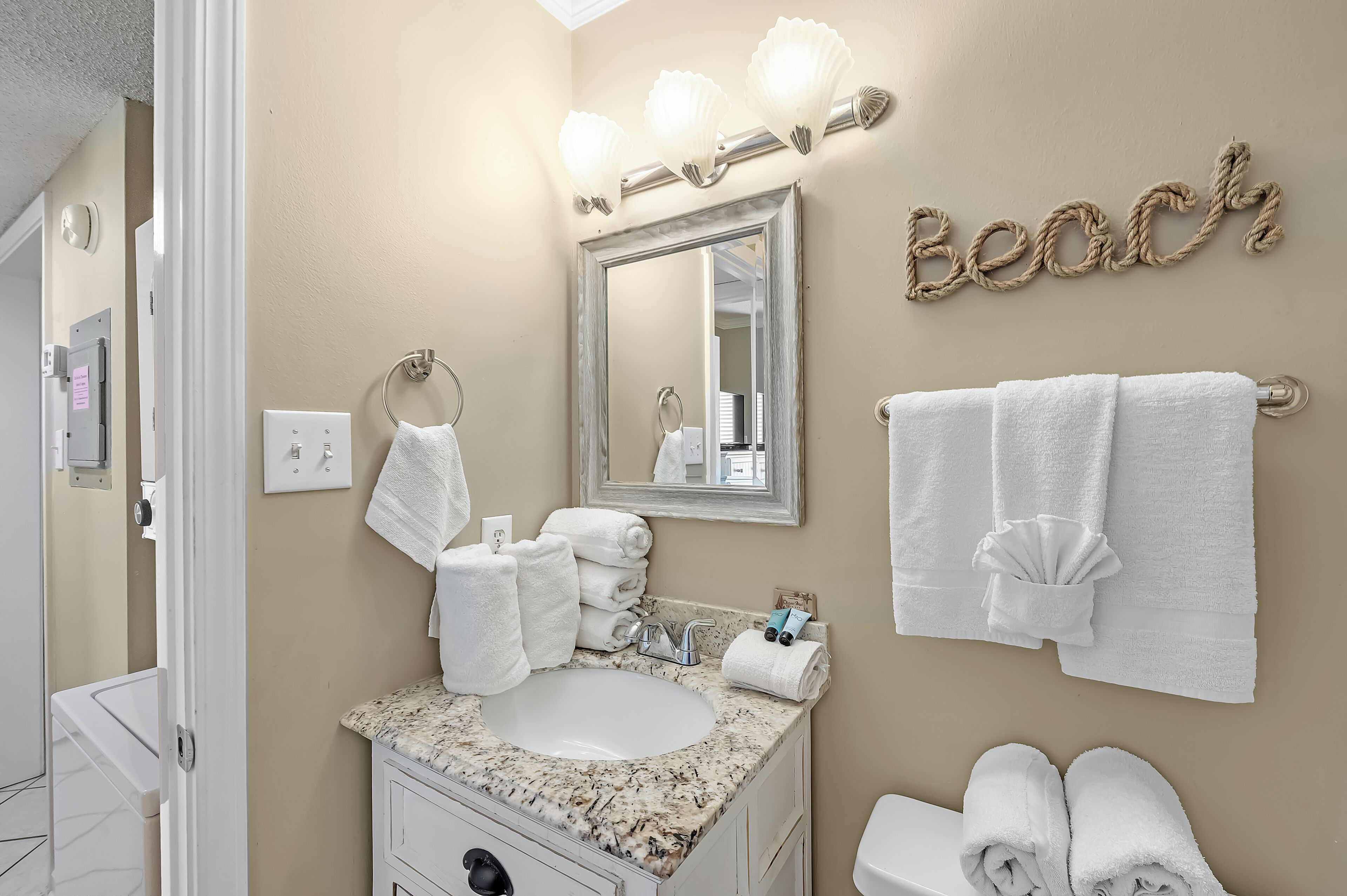 A bathroom with a sink, mirror, towel rack, and a wall with the word 'Beach' spelled out in rope.
