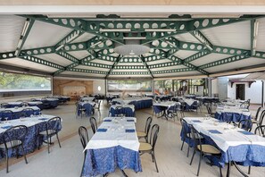 Un espace de restauration intérieur avec des tables disposées pour accueillir plusieurs personnes, un plafond haut avec des poutres apparentes de couleur verte et une vue sur l’extérieur.