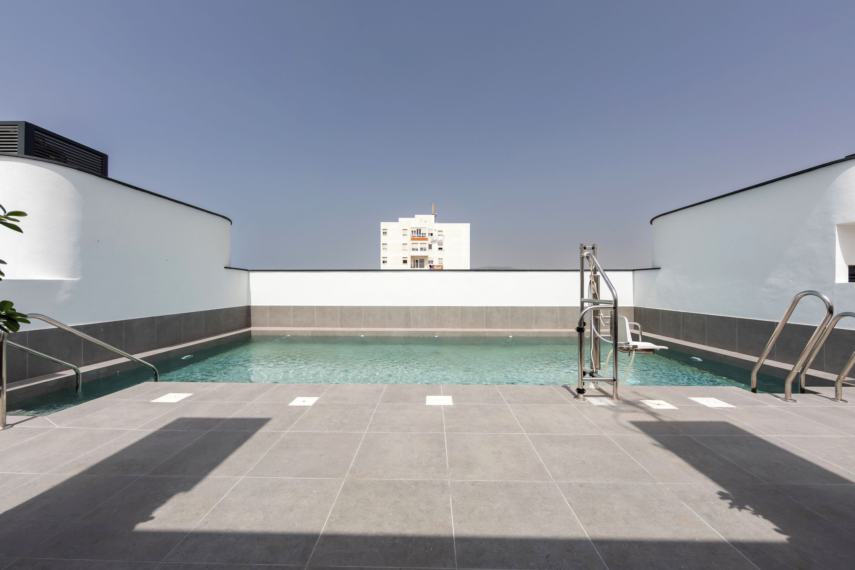 Una piscina en la azotea con agua azul cristalina, rodeada de baldosas grises y un edificio blanco al fondo.