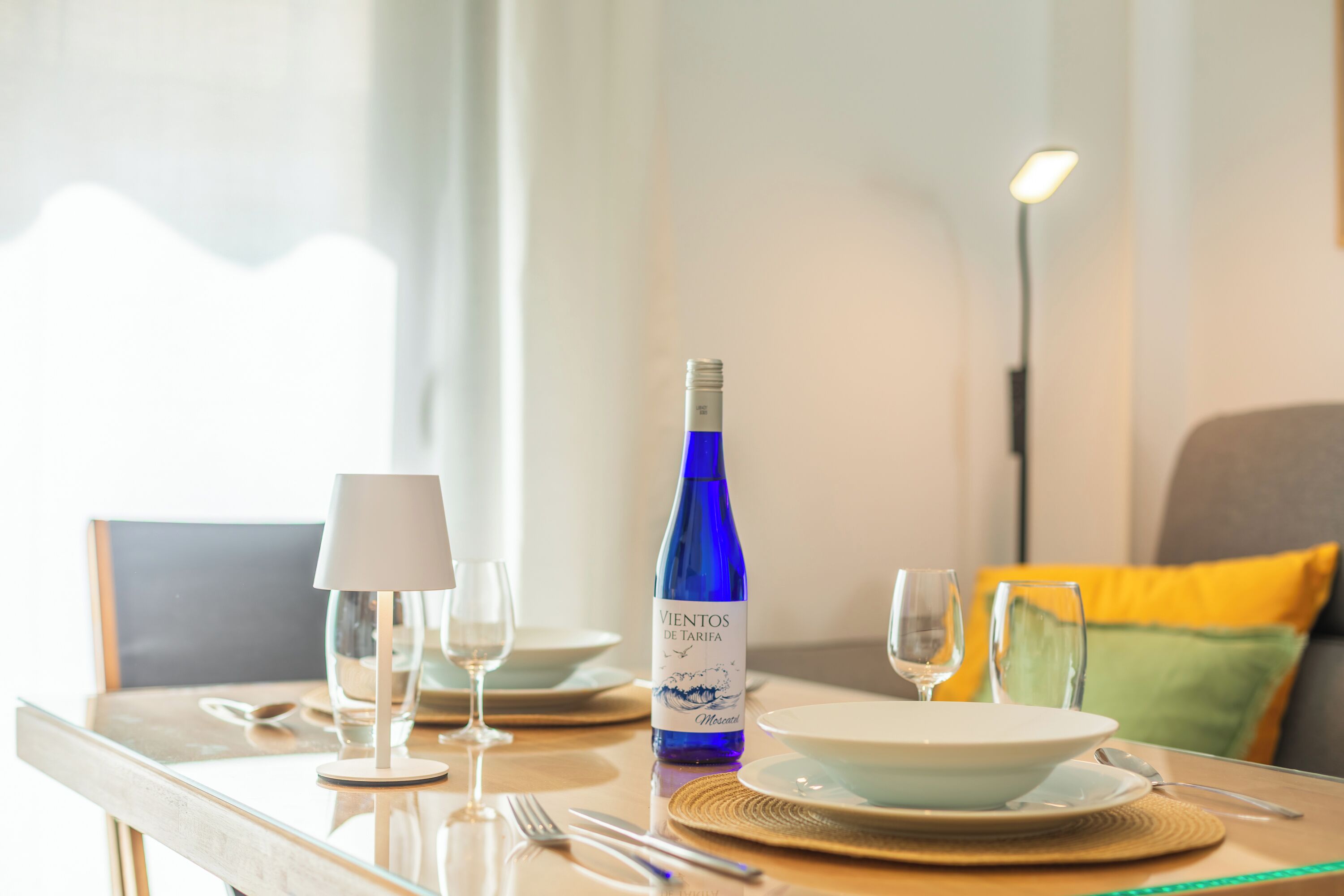 A dining table set with plates, cutlery, a wine bottle, and glasses, with a lamp and a floor lamp in the background.