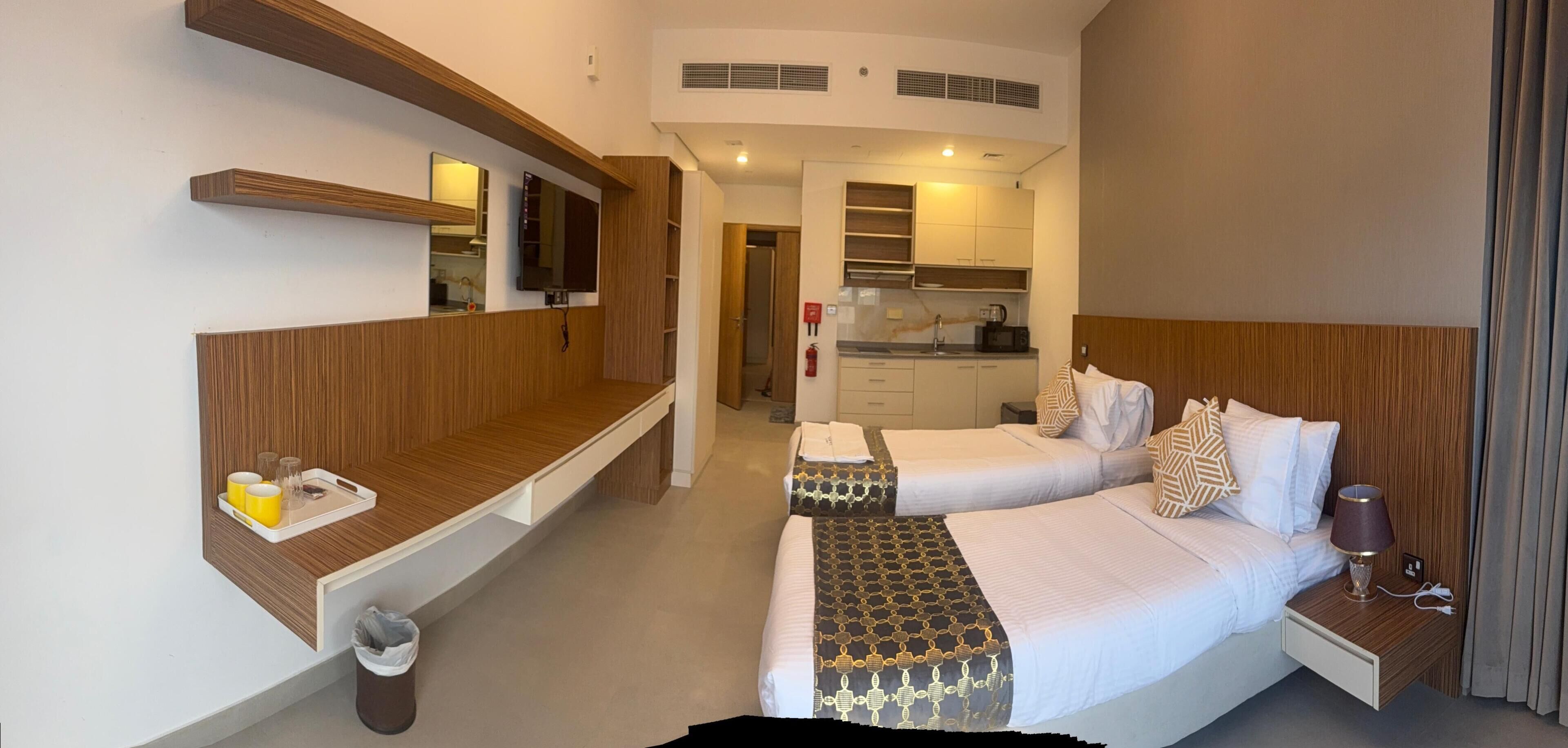 Een hotelkamer met een houten wastafel, een bed met beddengoed met een patroon, een nachtkastje met een lamp, en een kitchenette op de achtergrond.