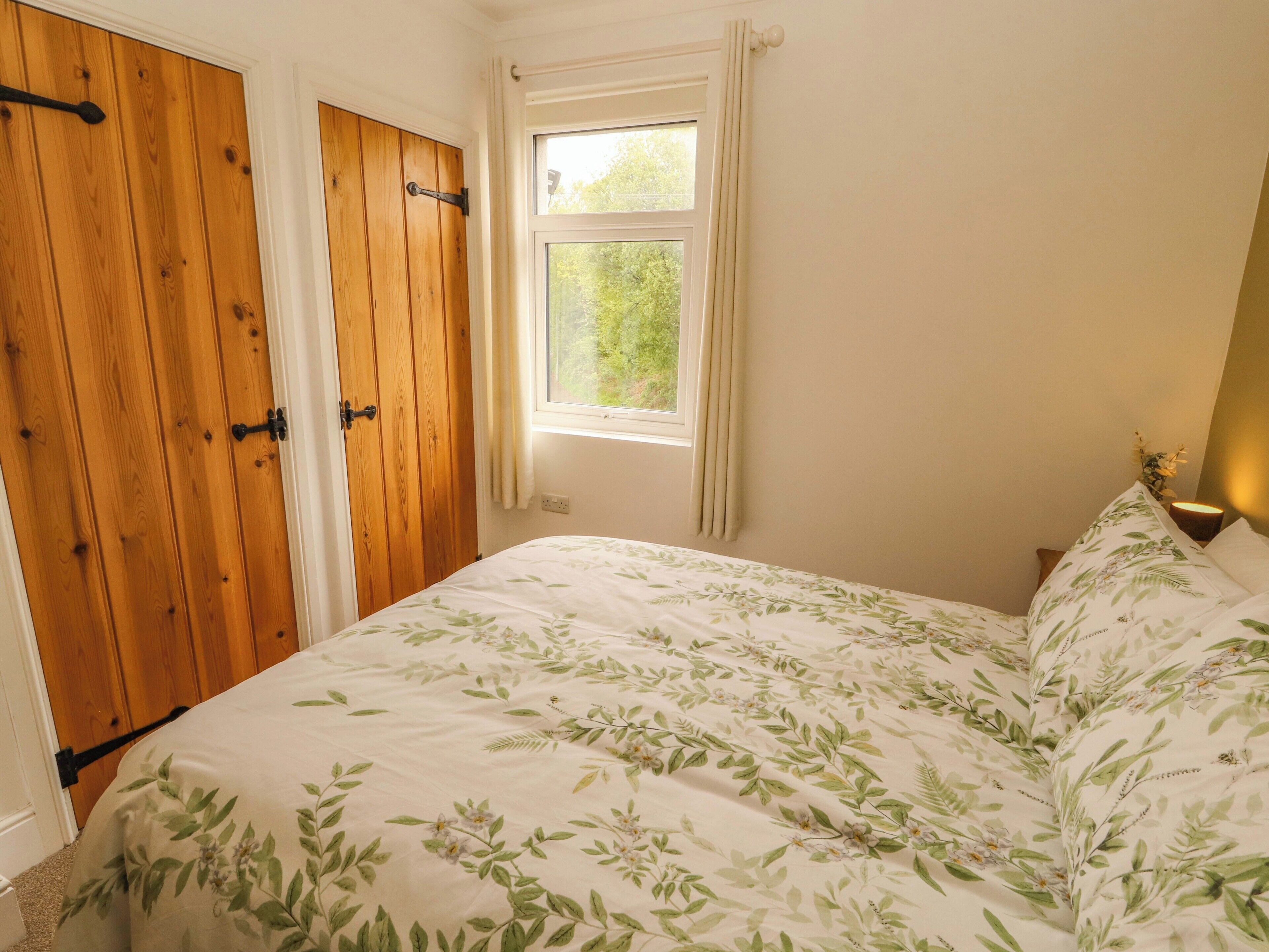 Un dormitorio con cama, armario de madera, ventana y mesita de noche con lámpara.