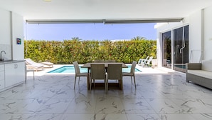A modern kitchen with white cabinets, a dining area with a table and chairs, a pool, and a view of a lush garden. - Family Villa With Private Pool in Sosua Ocean Village (Sosúa)