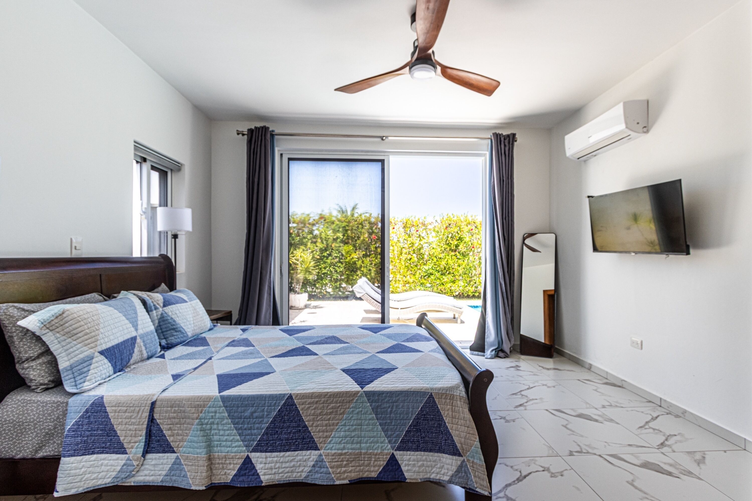 A bedroom with a bed, a ceiling fan, a TV, and a view of the outdoors.