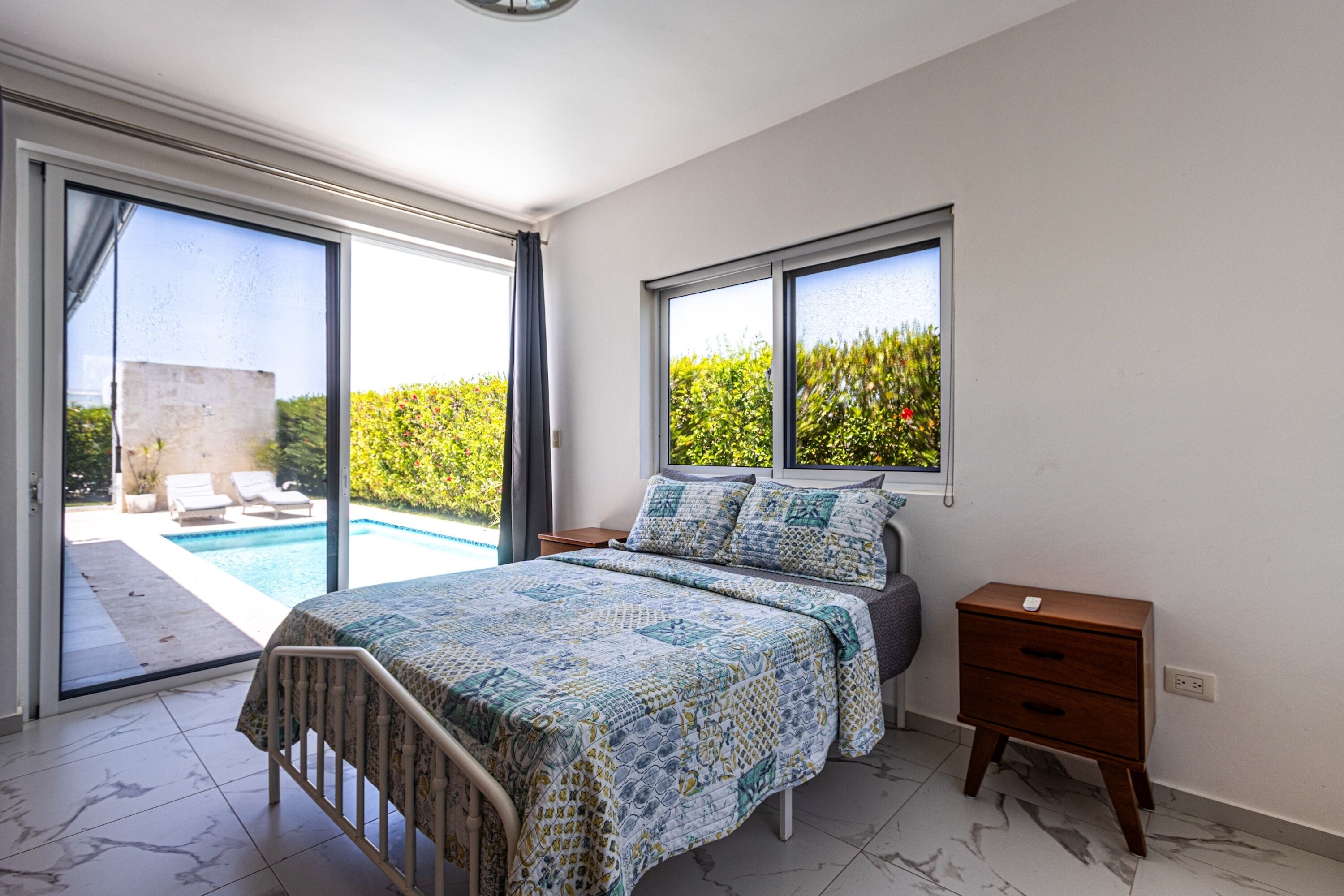 A bedroom with a bed, a nightstand, a window, and a view of a swimming pool.