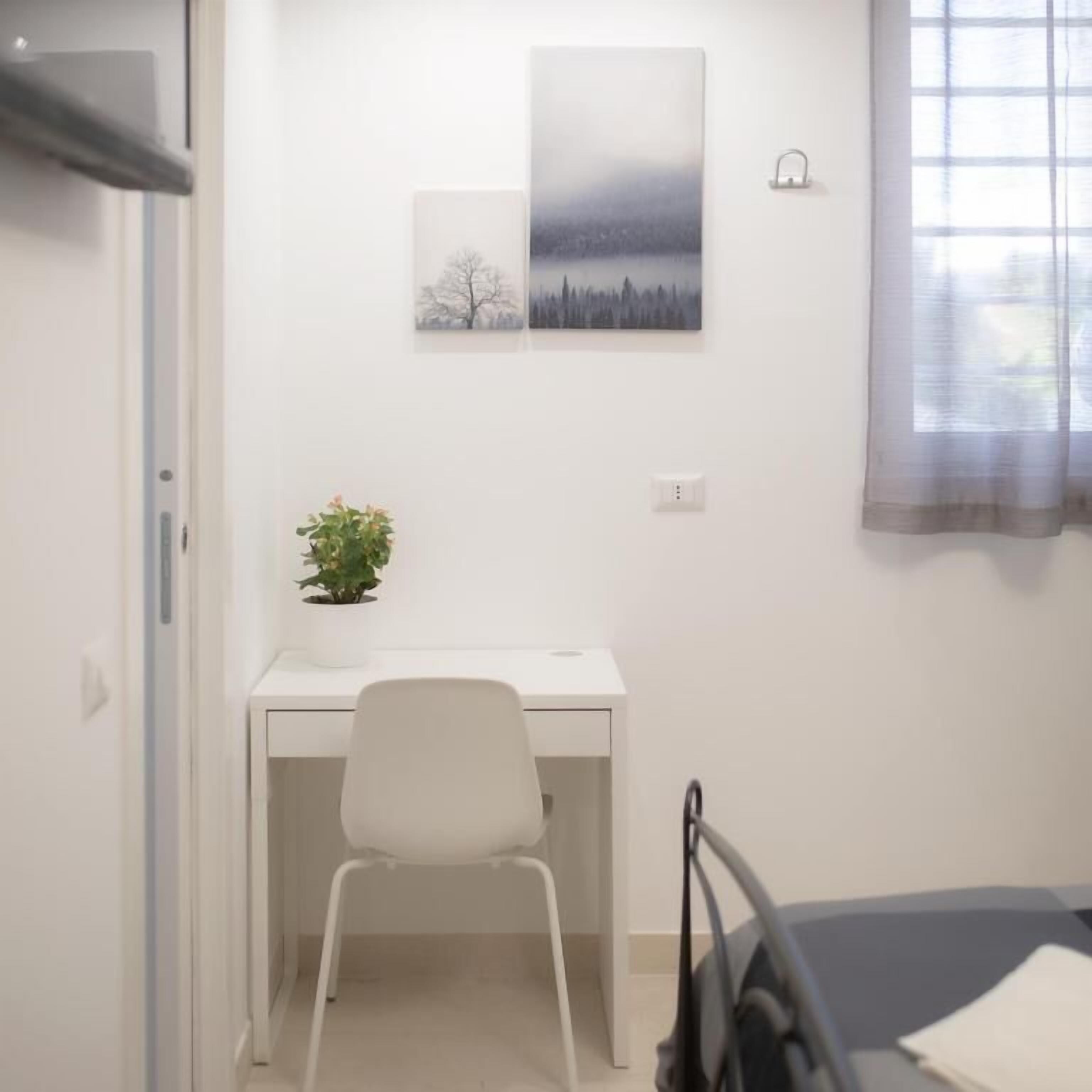 Een slaapkamer met een bed, een bureau met stoel, een bloempot en twee aan de muur bevestigde schilderijen.