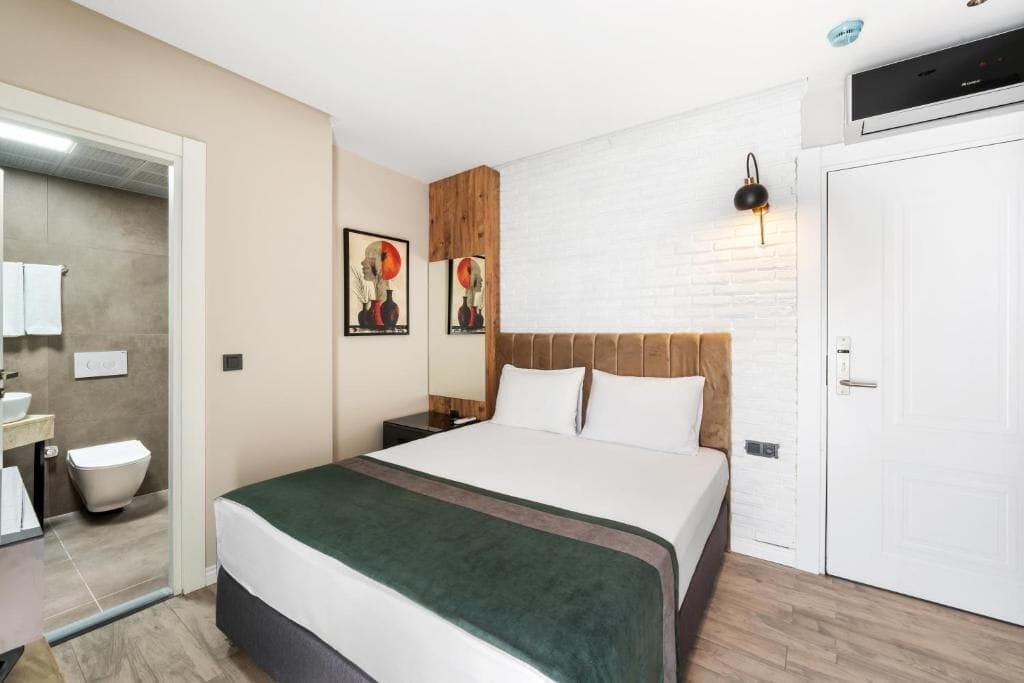 Een hotelkamer met een bed, een nachtkastje, een wandlamp, een deur en een witte bakstenen muur.