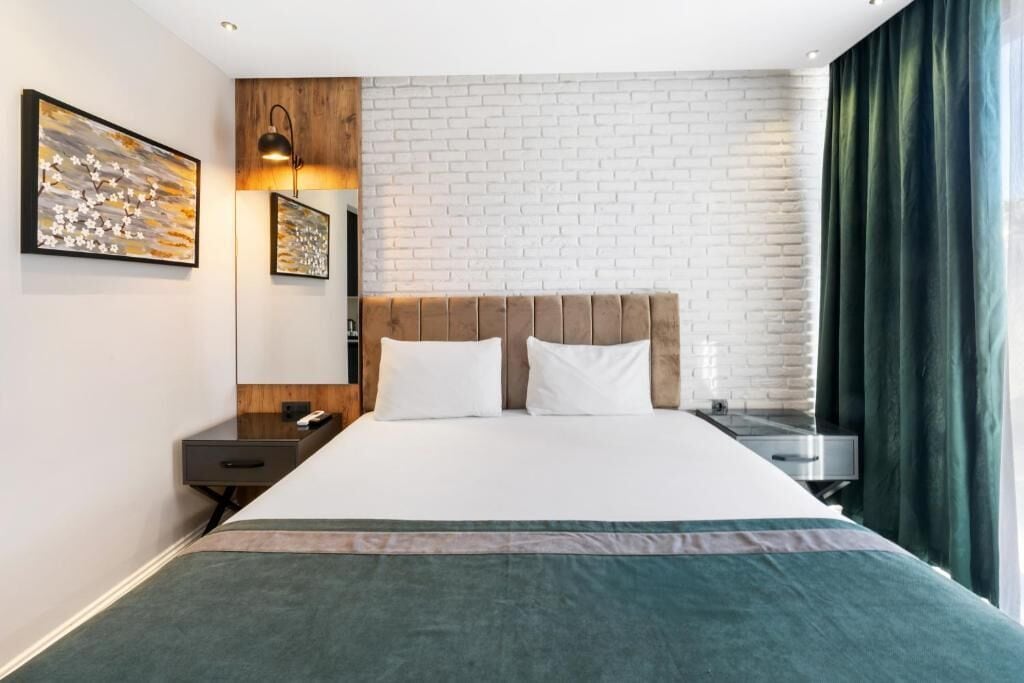 A hotel room with a brick-patterned wall, a bed with a headboard, a bedside table, a lamp, and a window with curtains.