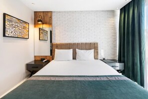 A hotel room with a brick-patterned wall, a bed with a headboard, a bedside table, a lamp, and a window with curtains. - Nivasole Lara (Antalya)