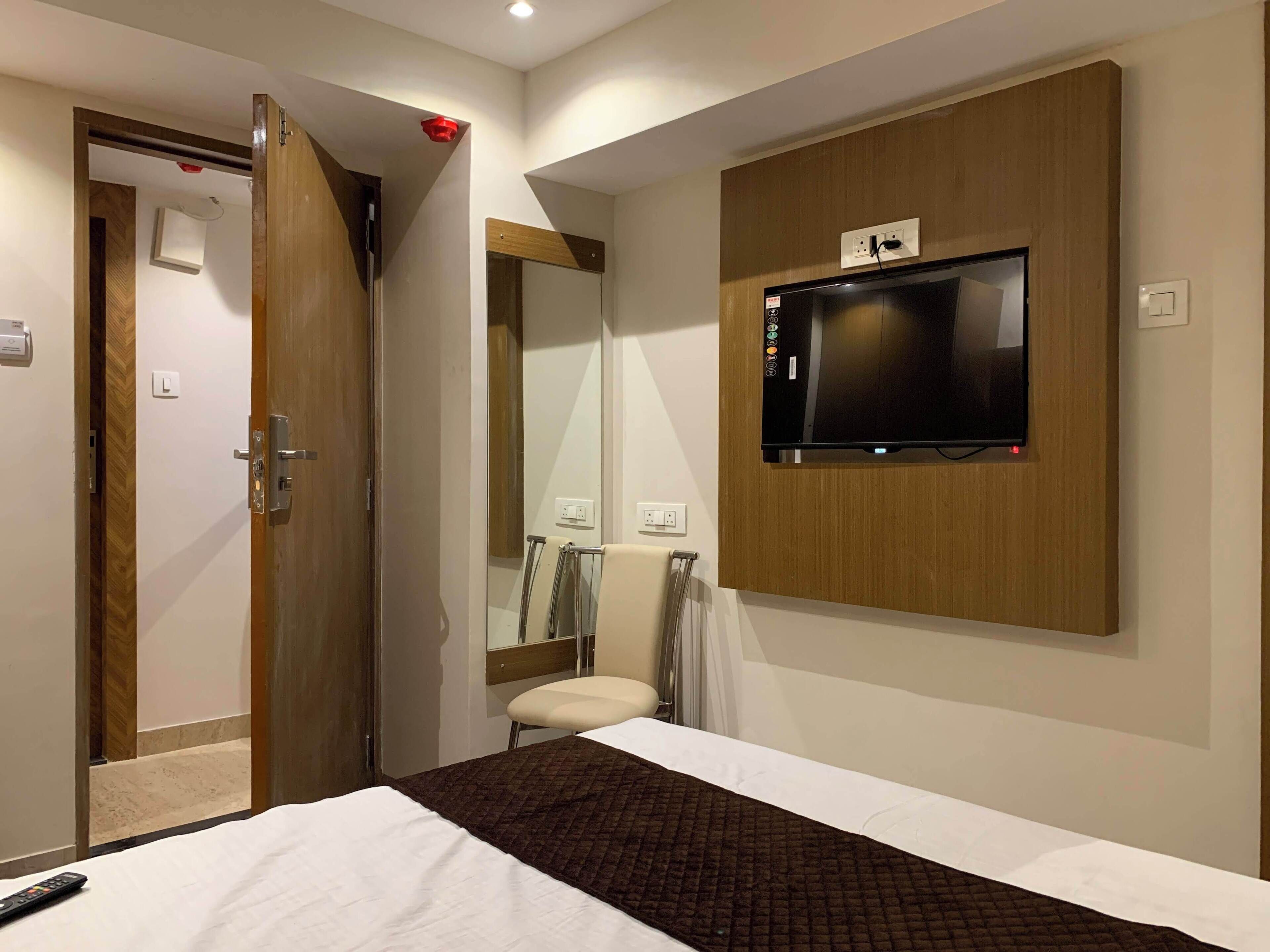 A hotel room with a bed, a TV mounted on the wall, a wooden door, and a bathroom visible through an open door.