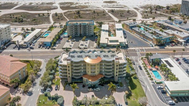 An aerial view of a coastal hotel with multiple buildings, a parking lot, and a beach in the background. - Crystal Palms 207 | Modern + Resort Amenities (Treasure Island)