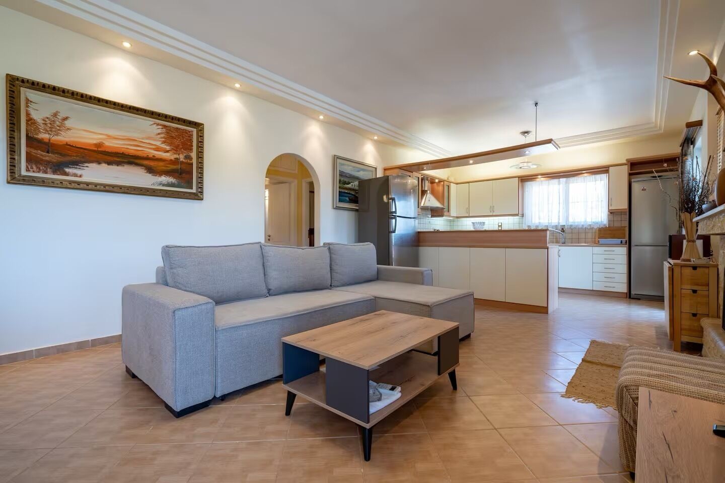 A living room with a grey sectional sofa, a wooden coffee table, and a kitchen area with white cabinets and a refrigerator.