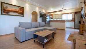 A living room with a grey sectional sofa, a wooden coffee table, and a kitchen area with white cabinets and a refrigerator. - Poolside Paradise Dilesi by ΜΡS (Πλάκα Δήλεσι)