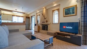 A modern living room with a sofa, coffee table, TV, fireplace, kitchen area, and a painting on the wall. - Poolside Paradise Dilesi by ΜΡS (Πλάκα Δήλεσι)