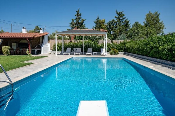 Ein rechteckiger Swimmingpool mit klarem, blauem Wasser, umgeben von einer gefliesten Liegefläche, einem überdachten Sitzbereich mit Stühlen und einem Gebäude aus Stein im Hintergrund.