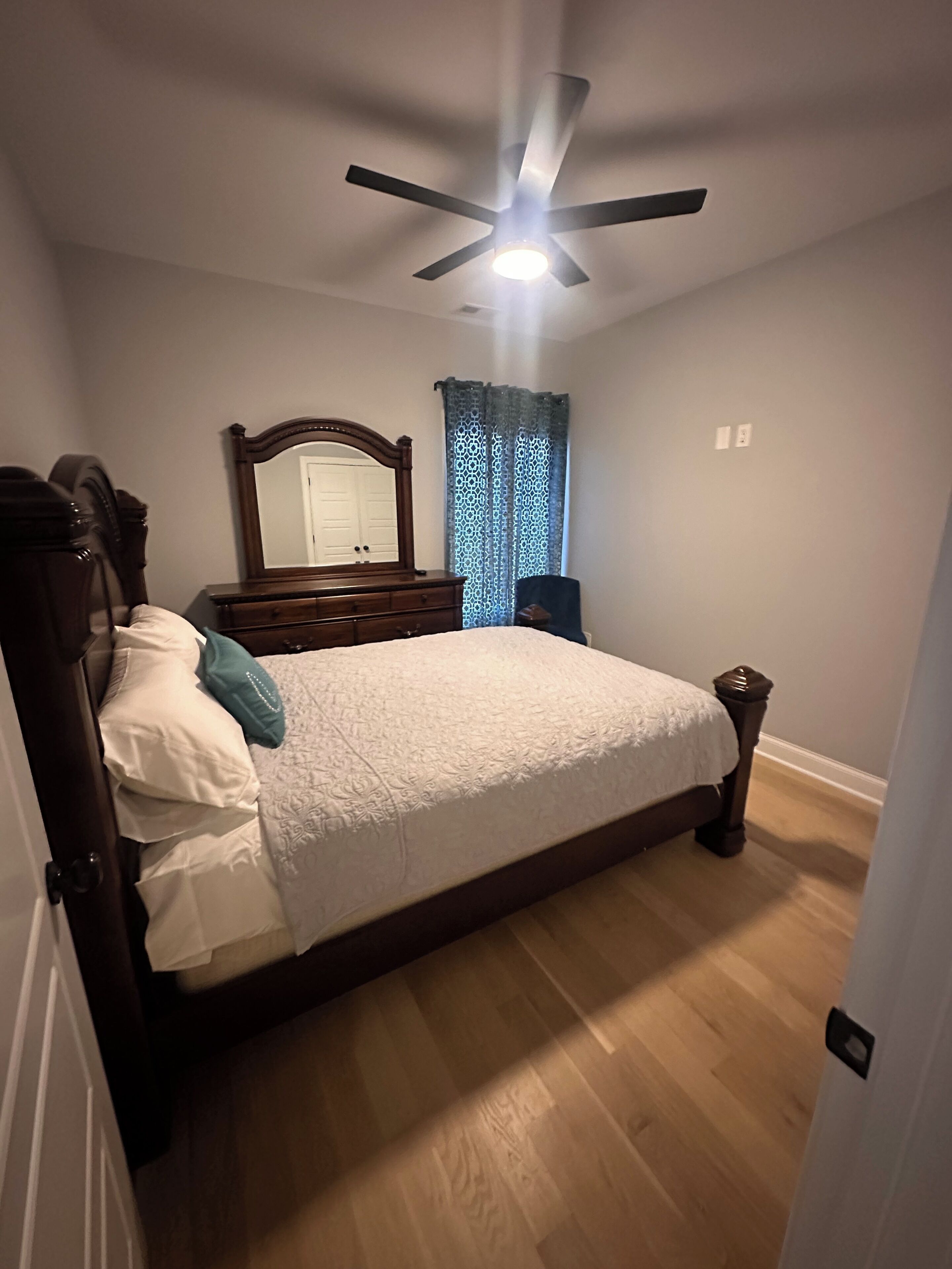 Quarto com cama, cômoda, espelho, ventilador de teto e janela com cortinas estampadas.