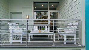 Ein Balkon mit weißen Stühlen und einem Tisch, einer Topfpflanze und einer Wandleuchte.