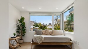 Ein Schlafzimmer mit einem Bett, einem großen Fenster, einer Zimmerpflanze und einem kleinen Tisch.