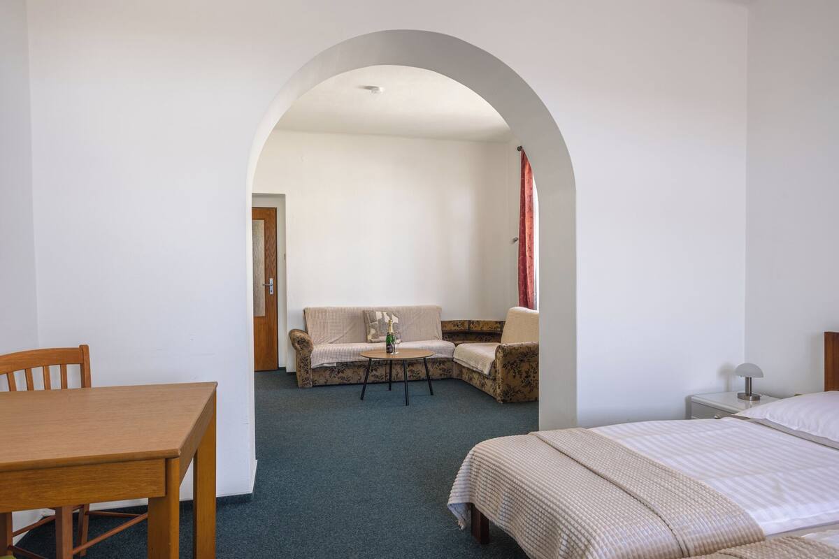 Ein Zimmer mit einem Holztisch, einer Couch, einem Bett und einem rundbogigen Durchgang zu einem anderen Raum.