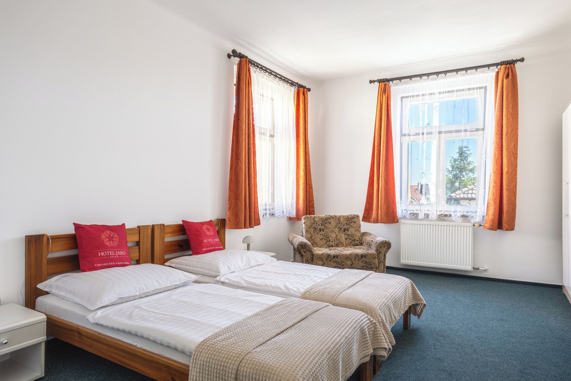 A hotel room with two beds, a chair, a radiator, and windows with curtains.