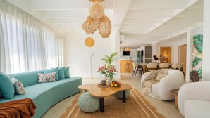 Una sala de estar moderna con sofá, mesa de centro y sillones. Se observa una cocina al fondo.
